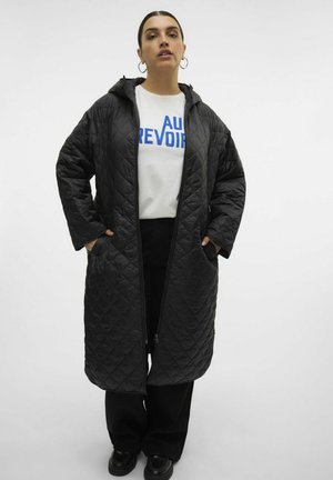 Manteau long noir matelassé avec capuche, doté d'une fermeture éclair avant et de grandes poches. Porte par-dessus un t-shirt blanc avec du texte bleu.