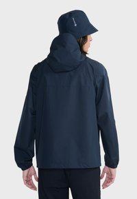 Laivaston vedenpitävä takki kiinnitettävällä hupulla ja joustavilla hihansuilla; siinä on ergonominen leikkaus ja sileä ulkopinta; mukana myös ämpärihattu.