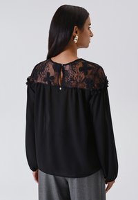 Una donna con lunghi capelli scuri indossa una blusa nera con un yoke in pizzo floreale trasparente e pantaloni grigi, mostrata di spalle contro uno sfondo semplice.