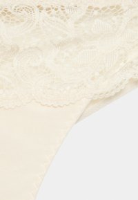 Thong de renda creme com padrões florais intrincados na cintura, feito de tecido de seda suave com uma textura lisa.