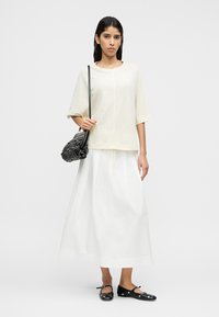 Λευκή βαμβακερή φούστα μέσης μήκους, συνδυασμένη με oversized πουκάμισο ανοιχτού κίτρινου χρώματος. Αξεσουάρ: μαύρη τσάντα και μαύρα επίπεδα παπούτσια με διακριτικές λεπτομέρειες.