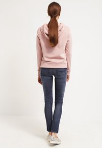 Kvinna med långt brunt hår i hästsvans, iklädd en rosa hoodie, mörkblå jeans och silverfärgade sneakers, står vänd bort mot en vit bakgrund.