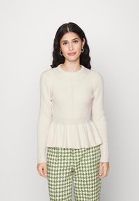 Pull en maille côtelée beige clair avec une silhouette peplum ; col rond et manches longues. Associé à un pantalon à motifs pied-de-poule vert et blanc.