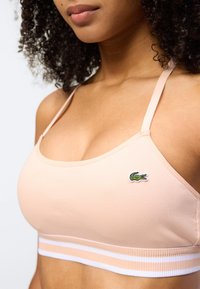 Γυναίκα που φοράει ροζ ρόμπα με λεπτές τιράντες και μικρό κεντημένο πράσινο λογότυπο κροκόδειλου στο στήθος.