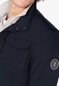 Chaqueta de color azul marino con cuello, que presenta cierres de presión metálicos y un gran bolsillo en el pecho. La manga izquierda tiene un parche con un logo redondo.