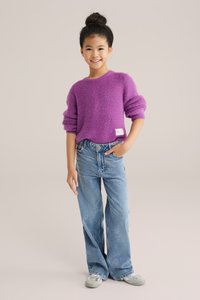 Lila bolyhos pulóver, bordás mandzsettákkal, világos kék, széles lábú farmernadrággal párosítva, amely díszített részletekkel rendelkezik. A modell mosollyal pózol.