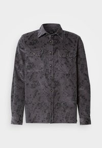 Chemise en côtes de velours gris avec un motif floral noir, deux poches sur la poitrine, un col pointu et des boutons métalliques. Tissu texturé avec des nervures verticales.