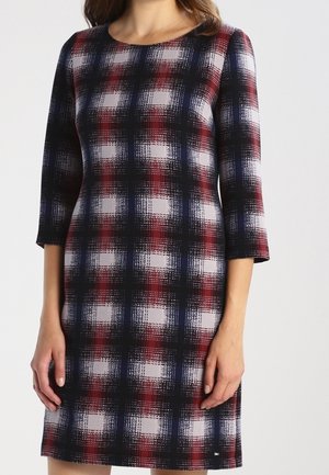 Femme portant une robe ajustée jusqu'aux genoux avec des manches trois-quarts et un motif à carreaux rouge, noir, blanc et bleu sur un fond clair.