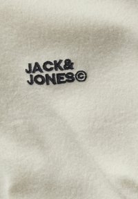Tessuto in pile crema spazzolato con logo ricamato "JACK & JONES" nero, caratterizzato da una morbida texture e un leggero splendore.