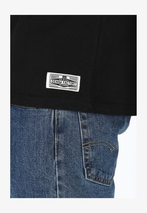 Schwarzes T-Shirt mit einem weißen "DENIM FACTORY"-Label nahe dem Saum, kombiniert mit blauen Jeans aus Denim mit gelben Nähten und einer Gesäßtasche.
