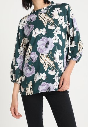 Blouse florale verte avec des fleurs violettes et blanches, col froncé et manches bouffantes trois-quarts, associée à un pantalon noir.