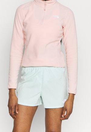 Nainen pukeutuneena vaaleanpunaiseen pitkähihainen neljännesvetoketjulliseen fleece-paitaan ja vaalean mintunvihreisiin shortseihin, joissa on joustava vyötärönauha.