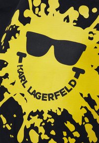 Żółte plamy farby z grafiką czarnych okularów przeciwsłonecznych i napisem "KARL LAGERFELD" ułożonym w łuk poniżej, na czarnym tle tkaniny.