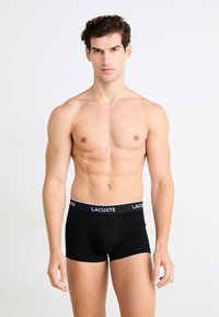 Zwarte boxershorts met een wit Lacoste-logo op de tailleband. De stof lijkt soepel en aangesloten, met een strakke en minimalistische uitstraling.