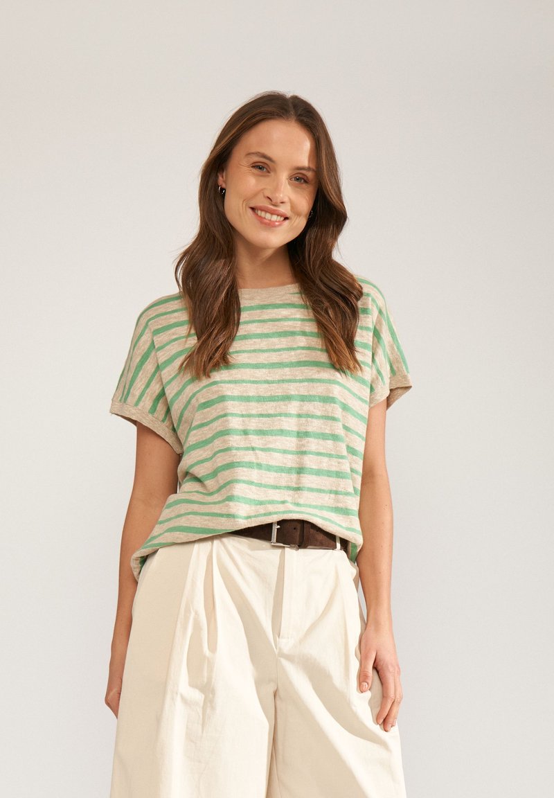 Femme souriante, portant un haut à manches courtes rayé vert et beige, rentré dans un pantalon crème taille haute avec une ceinture foncée.