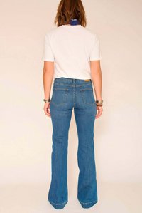 Jean en denim bleu évasé avec une taille haute, doté de poches arrière et d'une étiquette cousue. Porté avec un t-shirt blanc uni.
