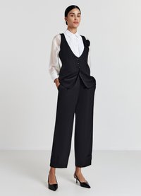 Gilet noir ajusté avec des boutons, superposé sur une chemise blanche plissée. Pantalons noirs à jambes larges présentant des ourlets repliés, associés à des escarpins noirs.