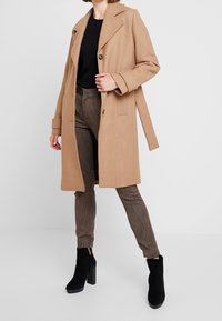 Manteau en mélange de laine couleur chameau avec col cranté et fermeture par boutons, associé à un pantalon marron texturé et des bottes noires à talons blocs.