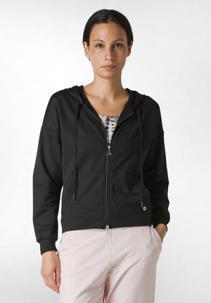 Black zip-up hoodie with a hood and drawstrings, featuring front pockets and a fitted design. Worn over a patterned shirt and light pants.