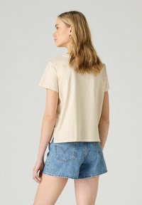 Beige puuvillat-paita lyhyillä hihoilla, jossa on halkiot helmassa, yhdistettynä sinisiin farkkushortseihin, nähtynä takaa.
