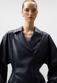 Donna con i capelli lisciati all'indietro, che indossa un giubbotto di pelle scura strutturato con ampi revers e orecchini a cerchio dorati, su uno sfondo chiaro e semplice.