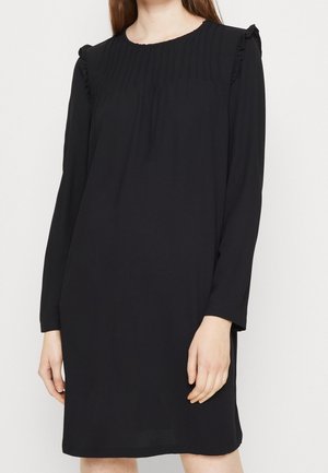 Woman wearing a long-sleeve black dress with pleated details and ruffled shoulders against a plain white background.