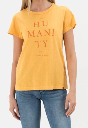 Femme portant un T-shirt jaune avec l'inscription « HUMANITÉ Inspirée par la nature », assorti d'un jean bleu, posant devant un fond clair.