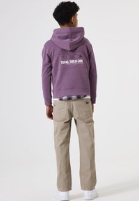 Un chico adolescente de espaldas, vestido con una sudadera morada con un diseño de montañas y texto, pantalones beige, zapatillas blancas y una camisa de cuadros debajo.