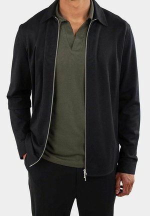 Mężczyzna w czarnej kurtce na zamek, pod którą ma oliwkową koszulkę polo, z rękami w kieszeniach na jednolitym tle.