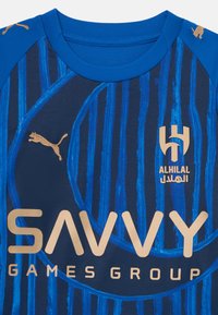 Tamno plavi sportski majica s plavim i zlatnim prugama, s Puma logotipom u zlatnoj boji, te natpisima "ALHILAL" i "SAVVY GAMES GROUP".