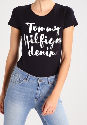 Czarna koszula z krótkim rękawem z białym napisem „Tommy Hilfiger denim”, noszona z jasnoniebieskimi dżinsami, ukazująca środkową część ciała i prawą rękę opartą na biodrze.