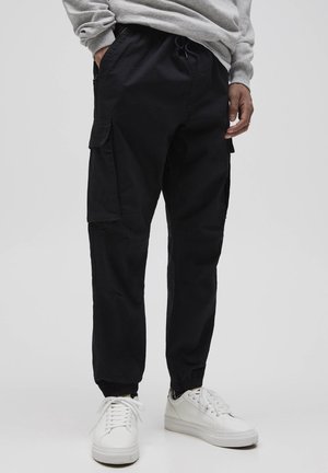 Homme portant un pantalon cargo noir avec des poignets élastiques, un sweatshirt gris et des baskets blanches, se tenant sur un fond uni.