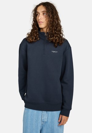 Navy blauwe zip-up sweatshirt met hoge kraag, zachte stof en klein wit logo op de borst. Gecombineerd met lichtblauwe, gestructureerde spijkerbroeken.