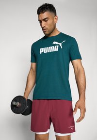 Teal kortærmet skjorte med stort hvidt "PUMA"-logo, parret med mørkerøde shorts med hvide detaljer. Person holder en håndvægt.