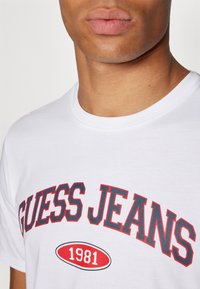 Balta medvilninė marškinėliai su apvaliu iškirpimu, ant kurių raudonai ir tamsiai mėlynai parašyta "GUESS JEANS", o žemiau teksto esanti raudona ovalo formos etiketė su užrašu "1981".