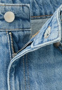 Gros plan sur un jean en denim bleu montrant une fermeture éclair en métal partiellement ouverte, un bouton avec gravure, et des détails de la ceinture cousue.