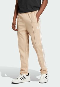 Beige track pants with white side stripes, elastic waistband, and pockets. Paired with white sneakers featuring black stripes.