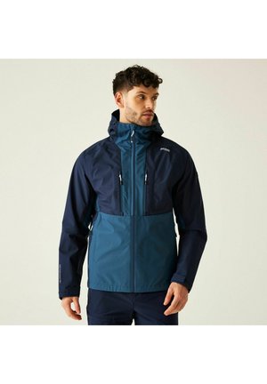 Mann mit kurzen lockigen Haaren, der eine zweifarbige, wasserfeste Kapuzenjacke in Marineblau und Blaugrün trägt und seitlich vor einem einfarbigen Hintergrund schaut.
