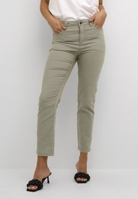 Mujer vestida con pantalones ajustados de oliva claro y sandalias de tacón trenzadas negras, de pie con las manos en los bolsillos frente a un fondo sencillo.