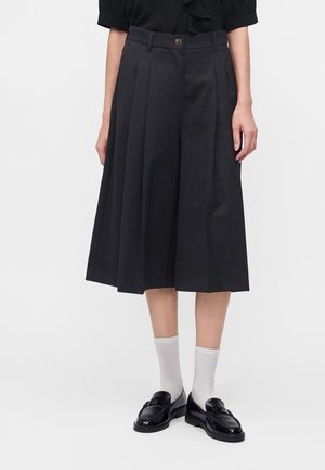 Donna che indossa una gonna midi nera plissettata, mocassini neri, calzini bianchi e una maglietta nera a maniche corte, in piedi su uno sfondo semplice.