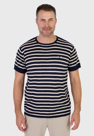 Man staat en draagt een navyblauw-wit gestreept shirt met korte mouwen en een klein blauw vogel-logo op de borst, en lichtgekleurde broek.