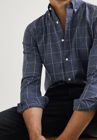 Marineblaue Hemdbluse mit weißem Karomuster, langärmlig und an den Ellenbogen hochgekrempelt, an einer sitzenden Figur mit schwarzer Hose.