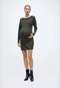 Εγκυμονούσα φόρεμα σε σκουρό γκρι πλεκτό ύφασμα, με μακριά μανίκια και άνετη εφαρμογή, συνδυασμένο με γυαλιστερά μαύρα μποτάκια αστραγάλου.