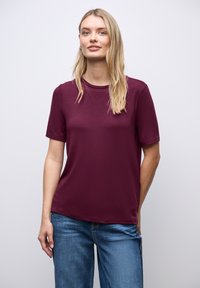 Kobieta o długich blond włosach, ubrana w bordową koszulkę z krótkim rękawem i niebieskie dżinsy, stojąca na tle jasnego, jednolitego tła.