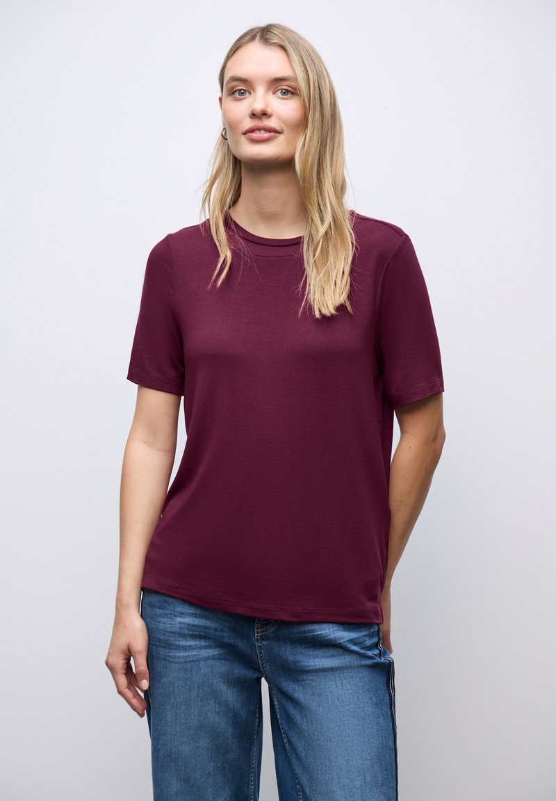 Kobieta o długich blond włosach, ubrana w bordową koszulkę z krótkim rękawem i niebieskie dżinsy, stojąca na tle jasnego, jednolitego tła.