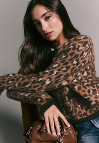 Leopardikuvioinen paita ruskeana ja mustana, pehmeällä tekstuurilla; mustalla resorilla varustettu helmassa. Etualalla pidettävä ruskea käsilaukku.