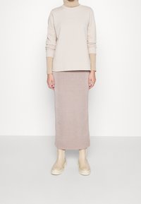 Donna vestita con un maglione beige a dolcevita, una gonna midi luccicante di un rosa chiaro e stivali chunky color crema, in piedi su uno sfondo chiaro e semplice.