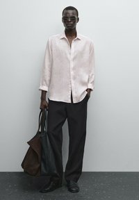 Light pink linen shirt, black trousers, and strappy sandals. Holds a black and brown tote bag. Clean lines, relaxed fit, minimal design.