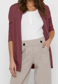 Cardigan bordeaux ouvert sur le devant, manches longues, porté sur un haut blanc à boutons et un pantalon beige taille haute avec deux boutons noirs.