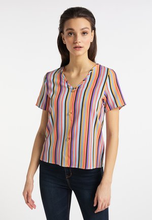 Junge Frau trägt eine kurzärmelige V-Ausschnitt-Bluse mit vertikalen mehrfarbigen Streifen und dunkle Jeans, steht vor einem schlichten Hintergrund.